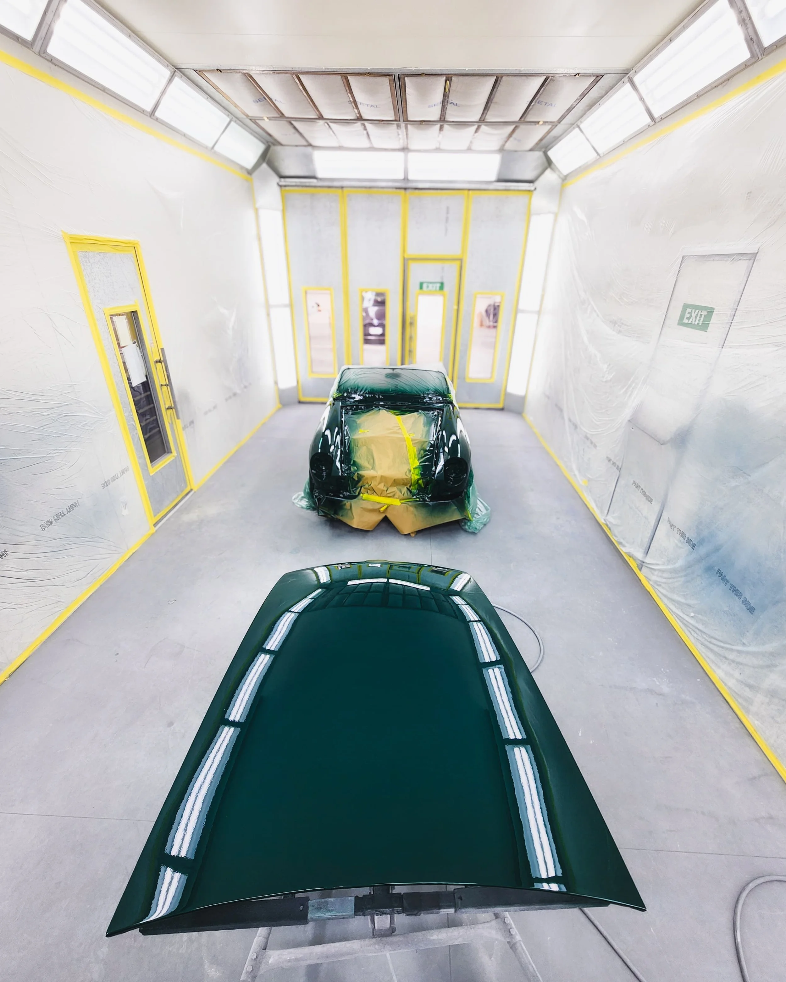 Car painted in a paint booth, with the car's hood and part of the front visible, and another car covered in plastic and masking tape for painting in the background.