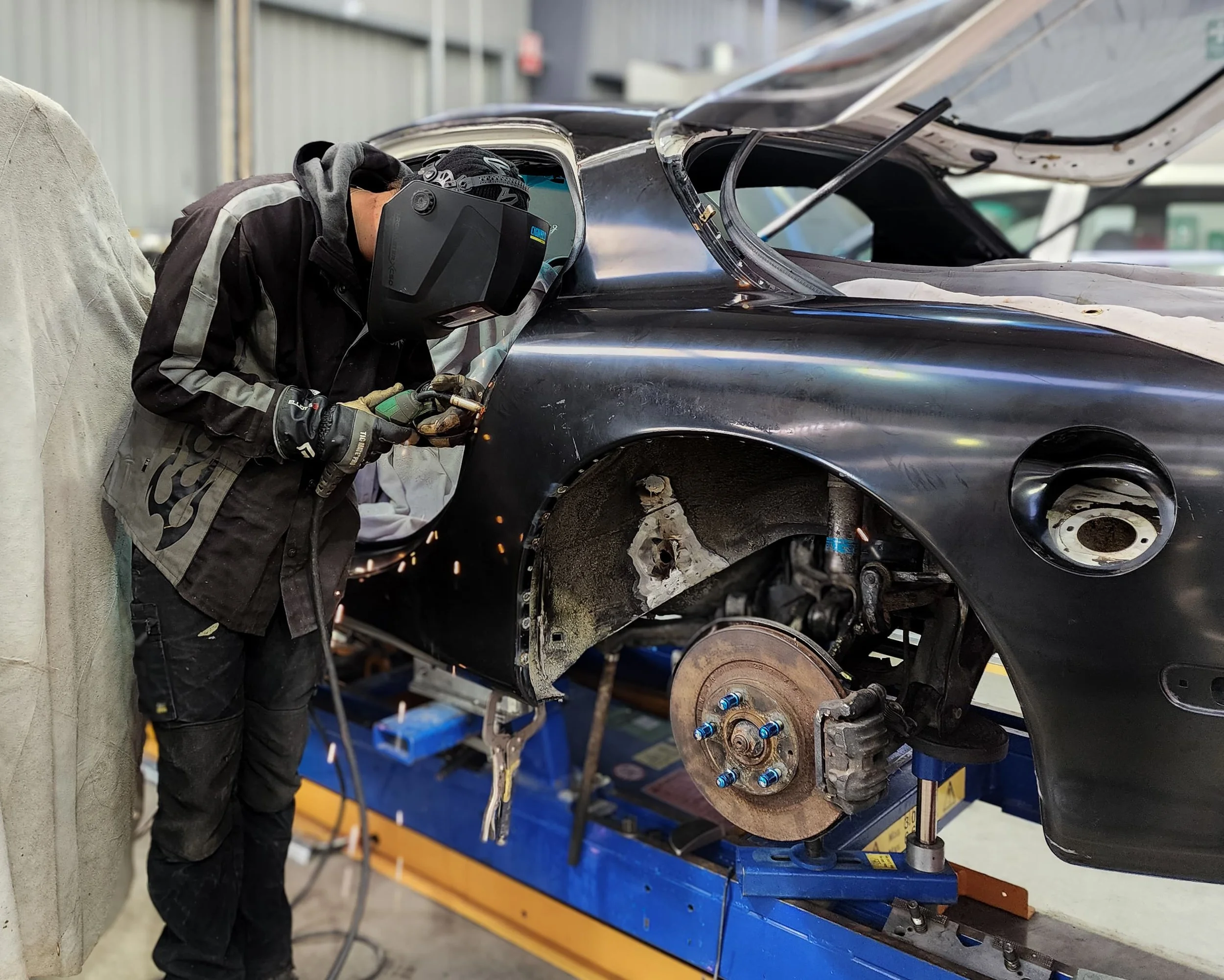 A mechanic welding a black car that is elevated on a blue hydraulic lift, with its front wheel removed in an auto repair shop.