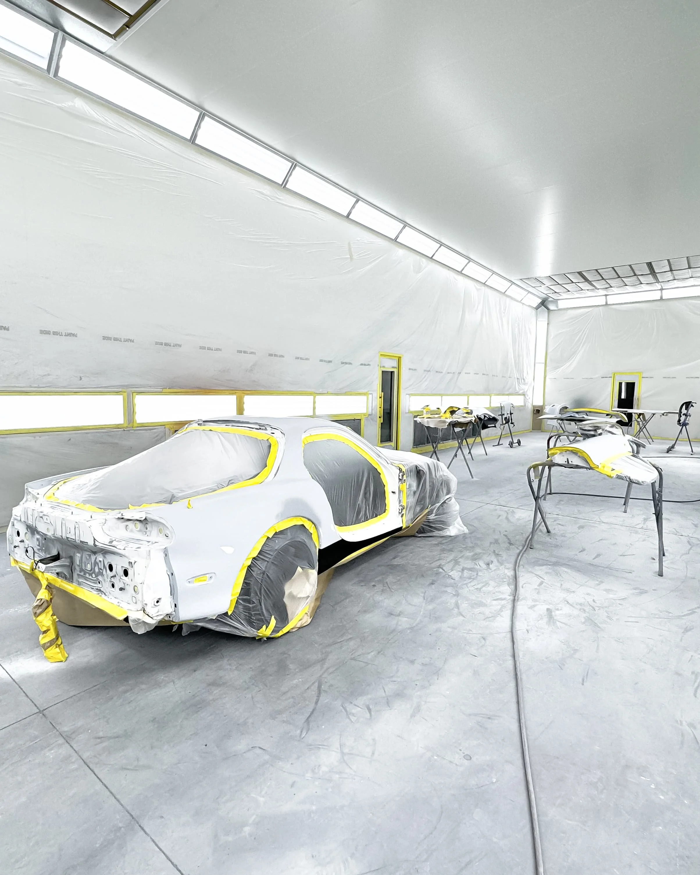 An automotive body shop with white painted car parts covered in plastic and masking tape, set up on stands and sawhorses, inside a well-lit spray booth with a white ceiling and walls.