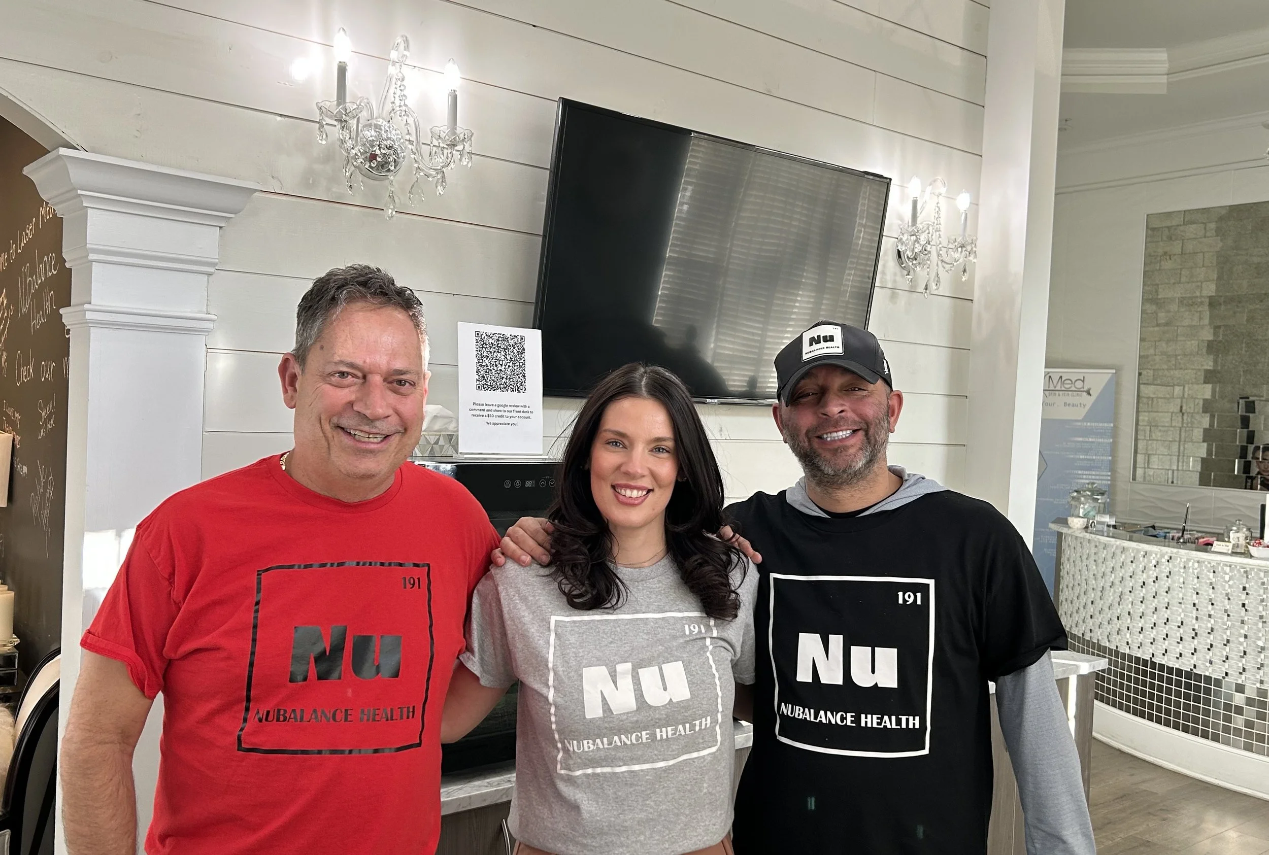 Three people smiling, wearing 'Nu Balance Health' shirts in a modern interior with a TV and chandeliers.