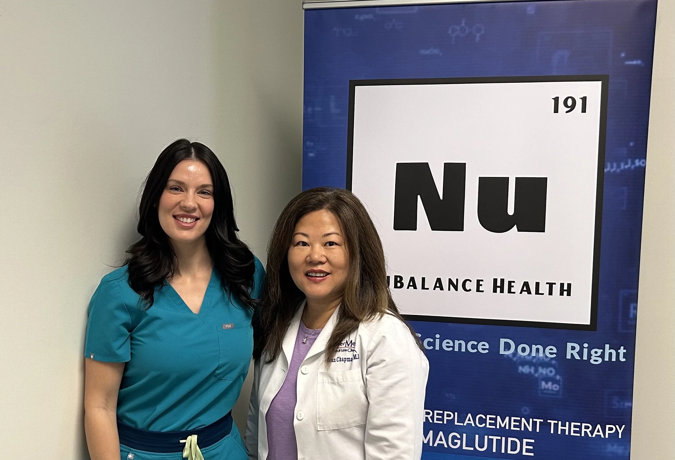 Two women posing in front of a medical banner, one in scrubs, the other in a white coat.