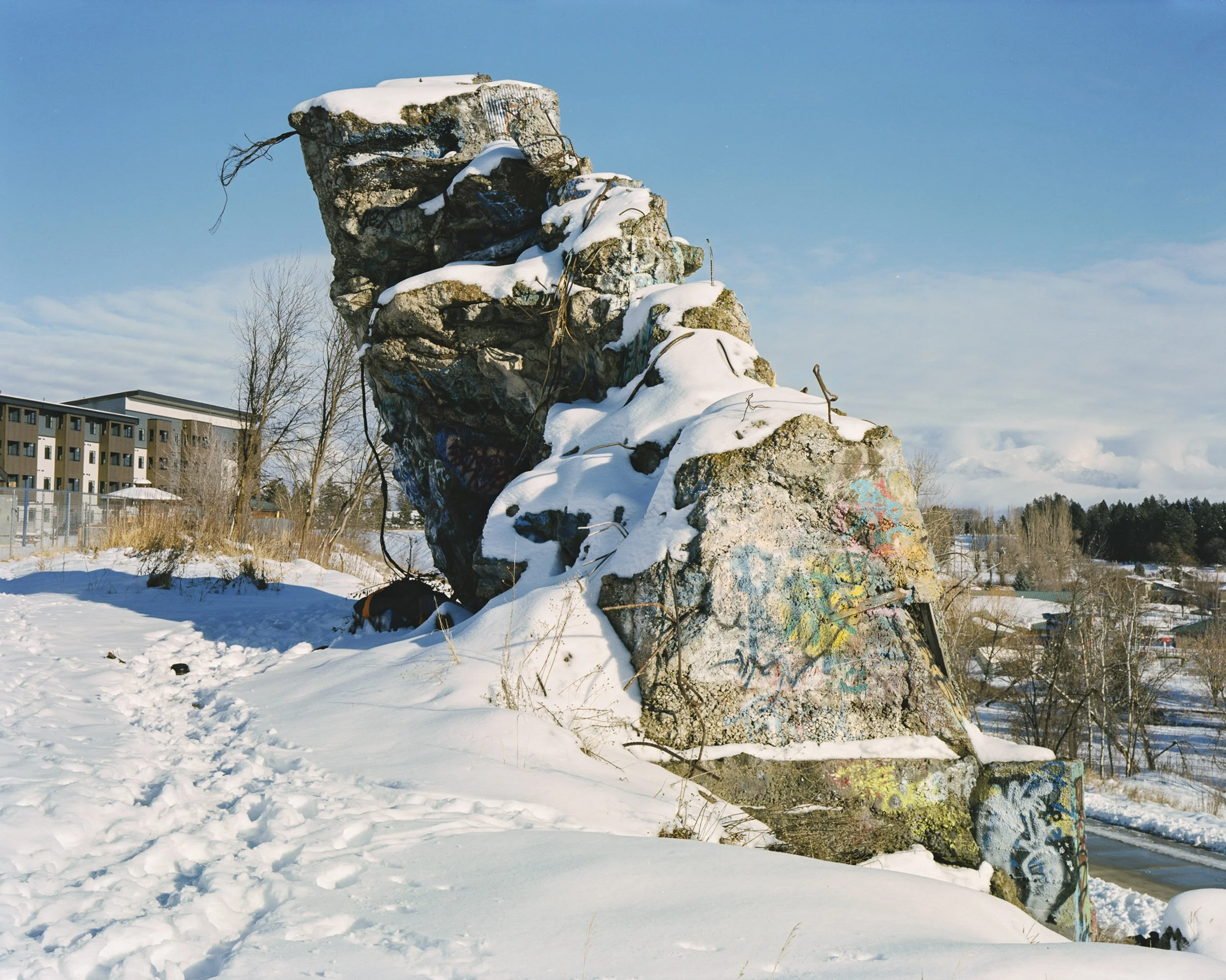 A sleeping bag and scattered belongings tucked behind an old bridge abutment overlooking the parkline trail and Woodland Park in Kalispell.