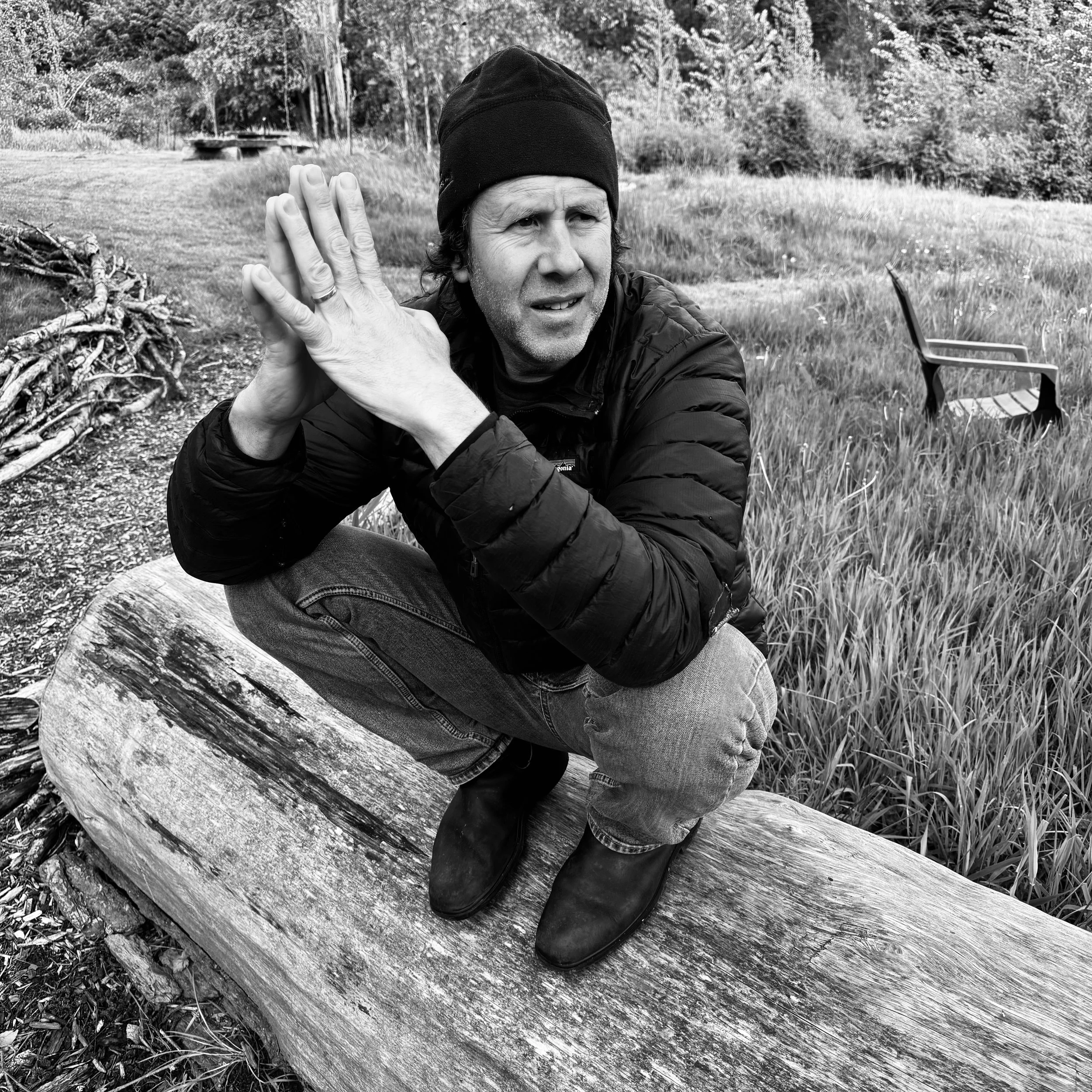 A man in a black beanie and jacket squatting on a log outdoors, looking to the side with his hands pressed together near his face, in a grassy area with a chair in the background.