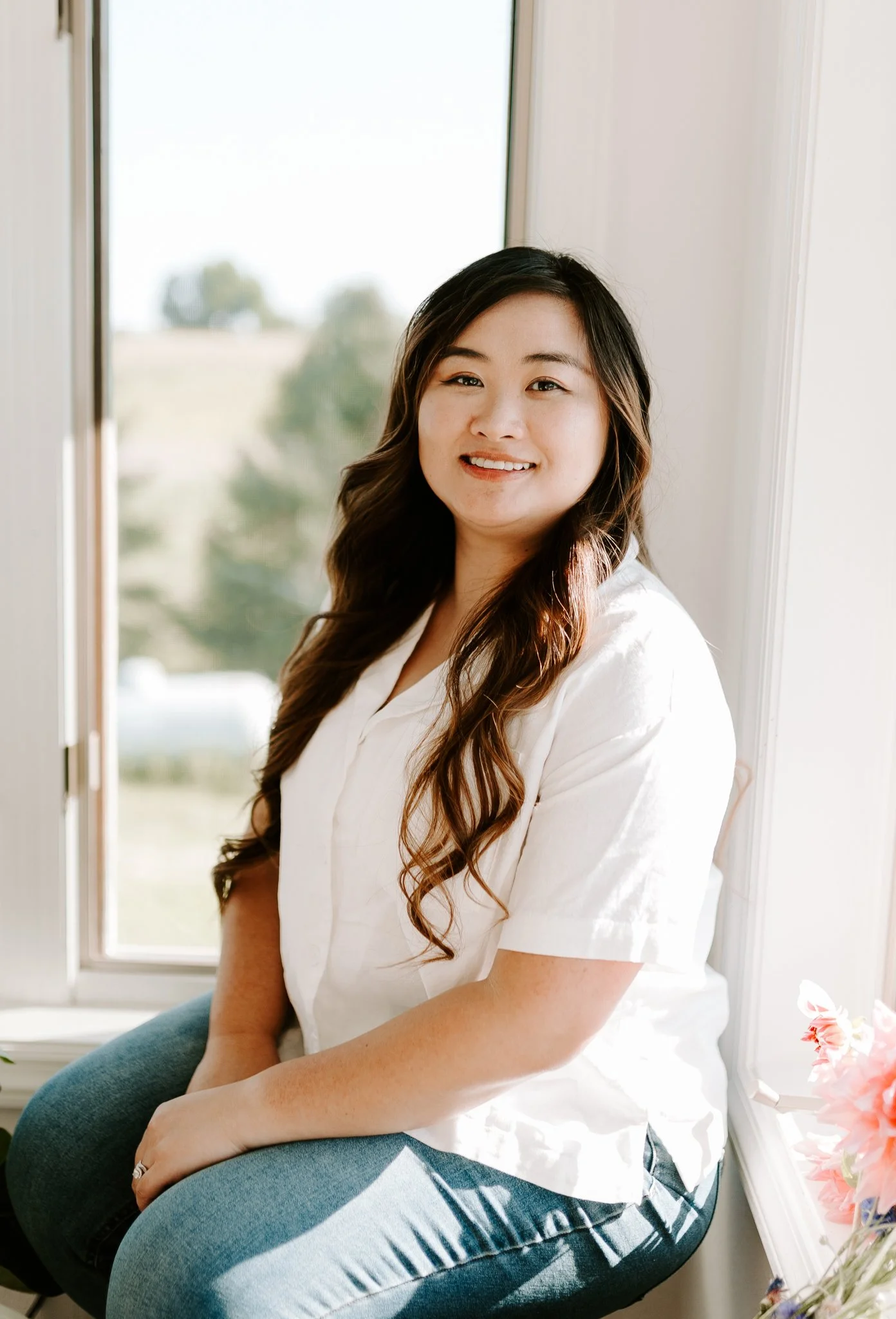 Person sitting on window ledge, smiling, wearing a white blouse and jeans, surrounded by flowers.
