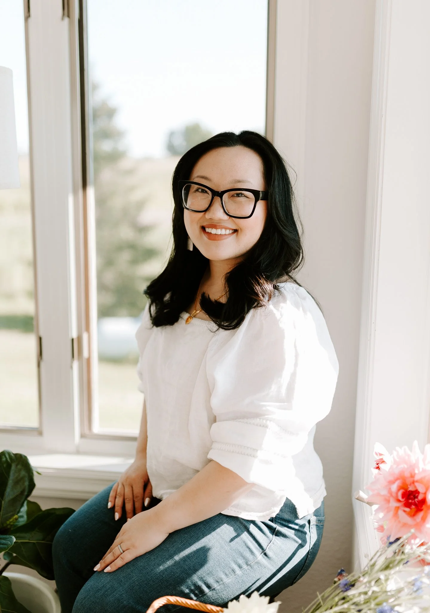 Woman in white blouse and glasses sitting near a window with flowers, smiling.