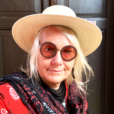 Woman wearing a wide-brimmed hat and round sunglasses, smiling, indoors with dark wood furniture.
