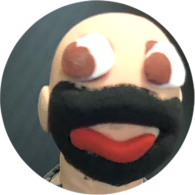 A puppet with a beige face, large white eyes with brown pupils, a black mustache, and a red tongue, against a dark background.