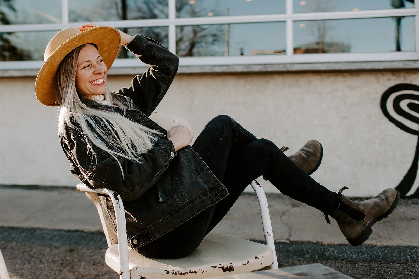 A smiling woman with long blonde hair wearing a wide-brimmed hat, black jacket, black pants, and brown boots, sitting on a white vintage chair outdoors, leaning back with her eyes closed.