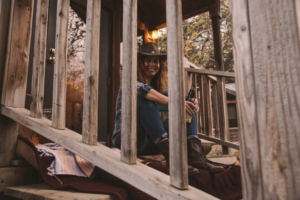 A woman sitting on a porch, smiling, holding a bottle, wearing a hat and casual clothing, surrounded by autumn trees.