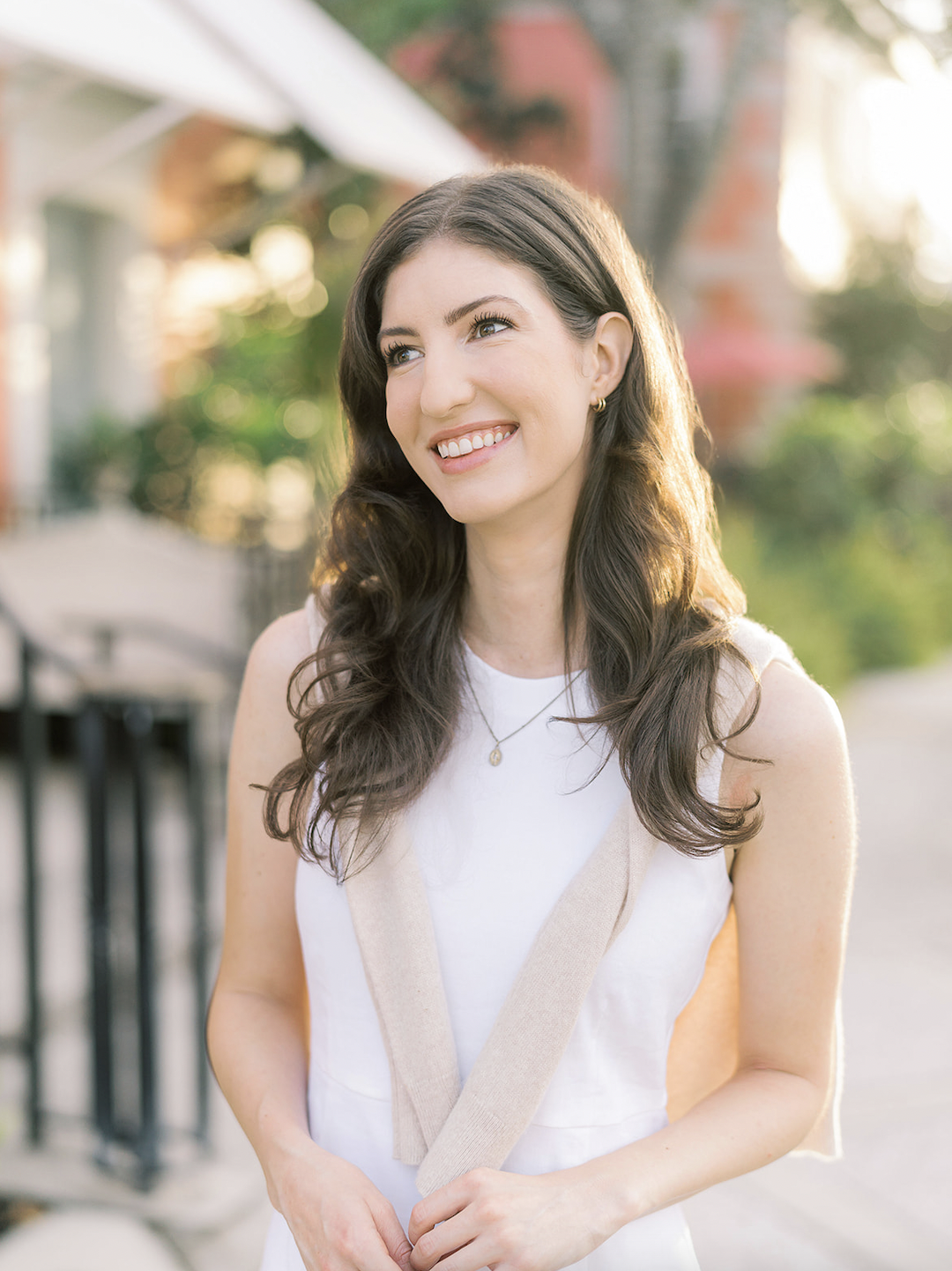 A smiling woman with long dark hair standing outdoors in natural sunlight, wearing a white sleeveless top, a beige cardigan draped over her shoulders, and a gold necklace, with a background of blurred greenery and buildings.