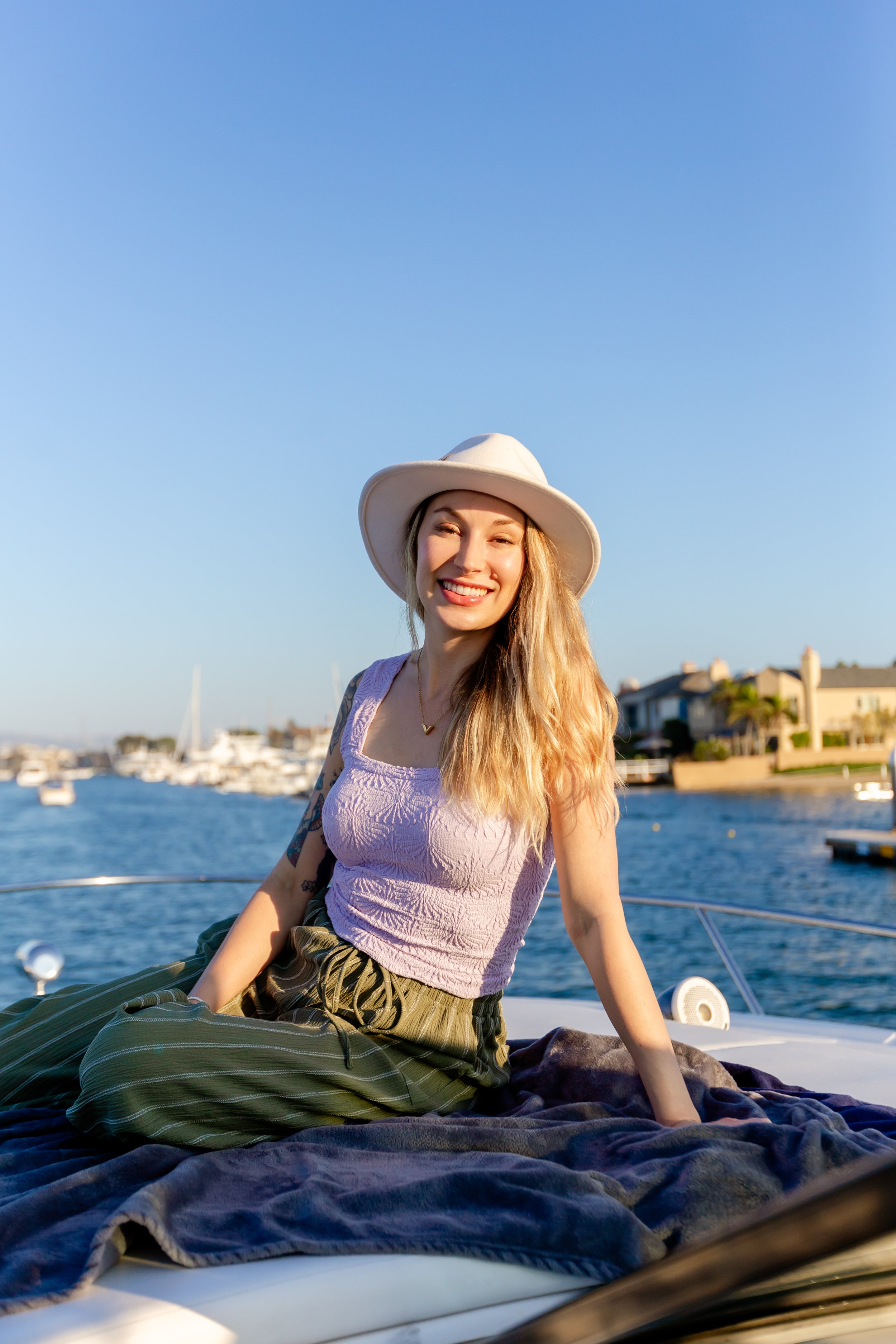 A smiling young woman sitting on a boat near the water, wearing a wide-brimmed hat, a light purple sleeveless top, and striped green pants, with boats and houses in the background under a clear blue sky.