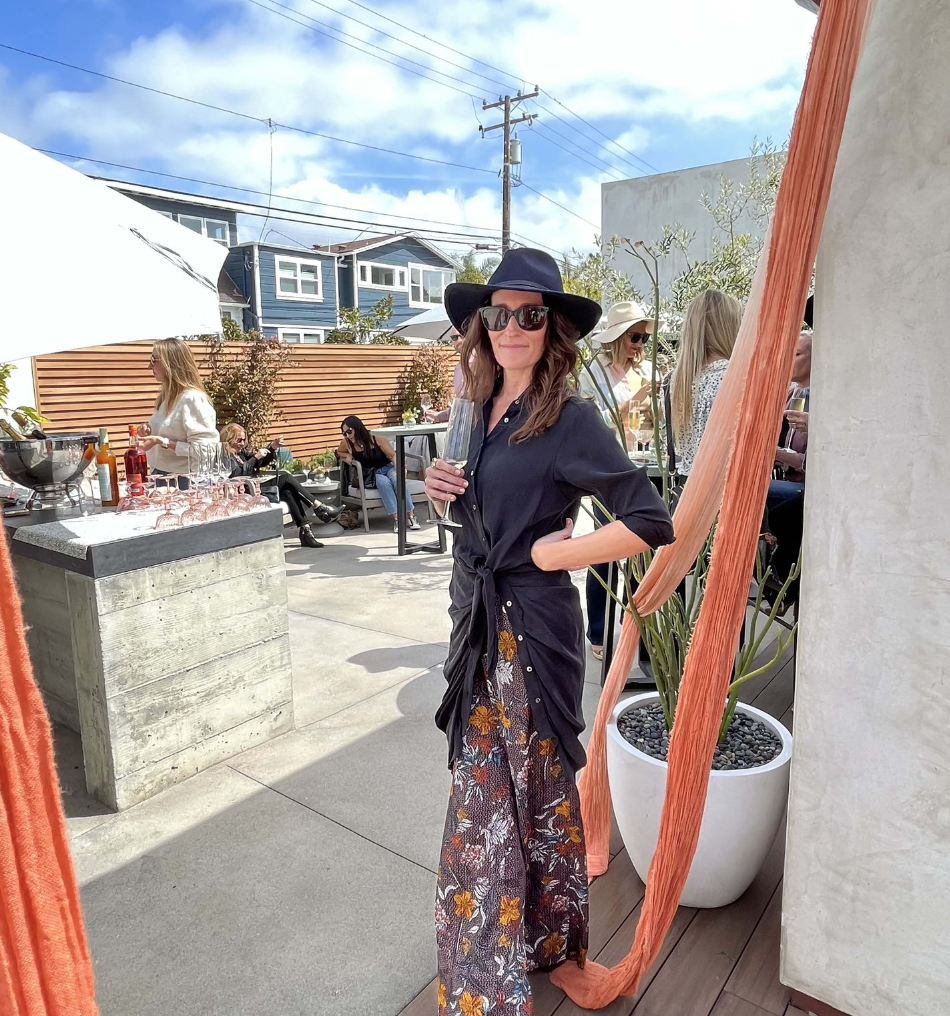 Woman wearing a black wide-brimmed hat, black sunglasses, and a dark shirt, standing outdoors at a social gathering. She is holding a drink and has a slight smile. In the background, there are other people, potted plants, and a wooden fence, with partly cloudy skies overhead.