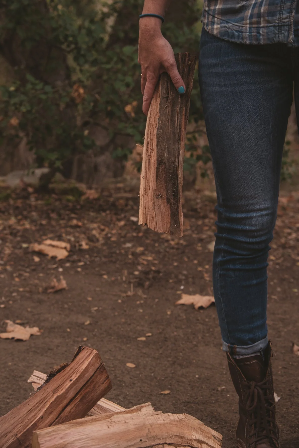 Person holding a piece of split firewood outdoors, with fallen logs on the ground and a background of green foliage.