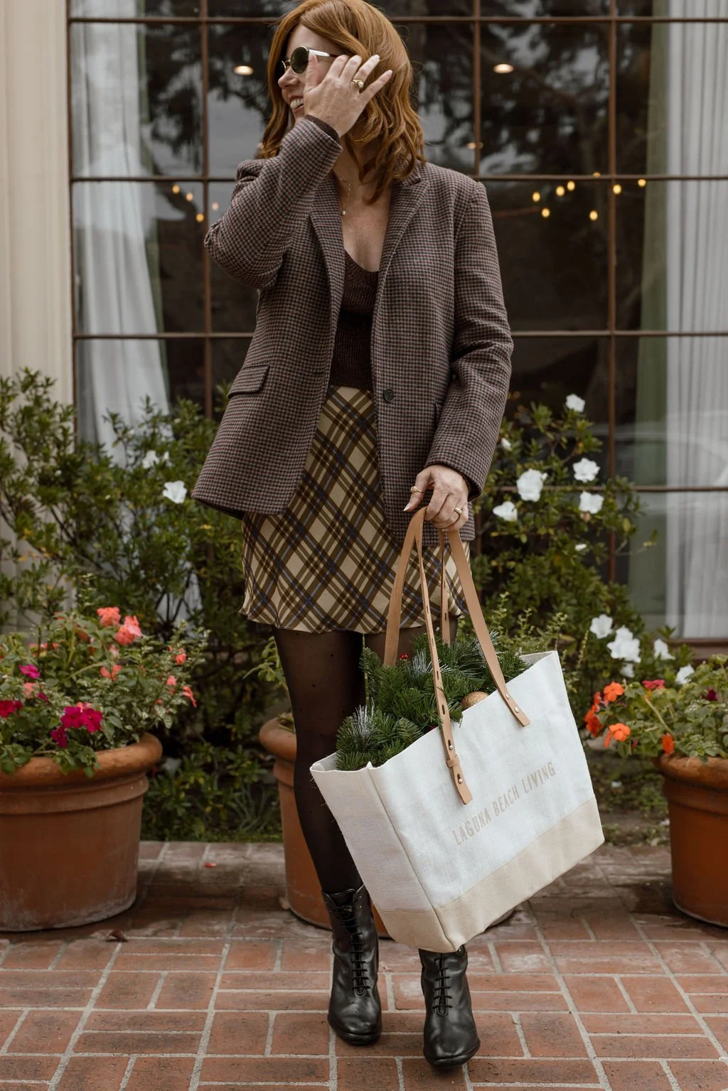 A woman in a brown plaid jacket, black tights, and black boots standing outdoors on a brick patio with potted flowers, holding a white tote bag with greenery inside, smiling and adjusting her sunglasses.