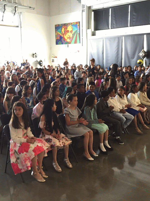 Group of young children sitting in chairs, attending a school event in a large indoor space with colorful artwork on the wall and balloons as decorations.