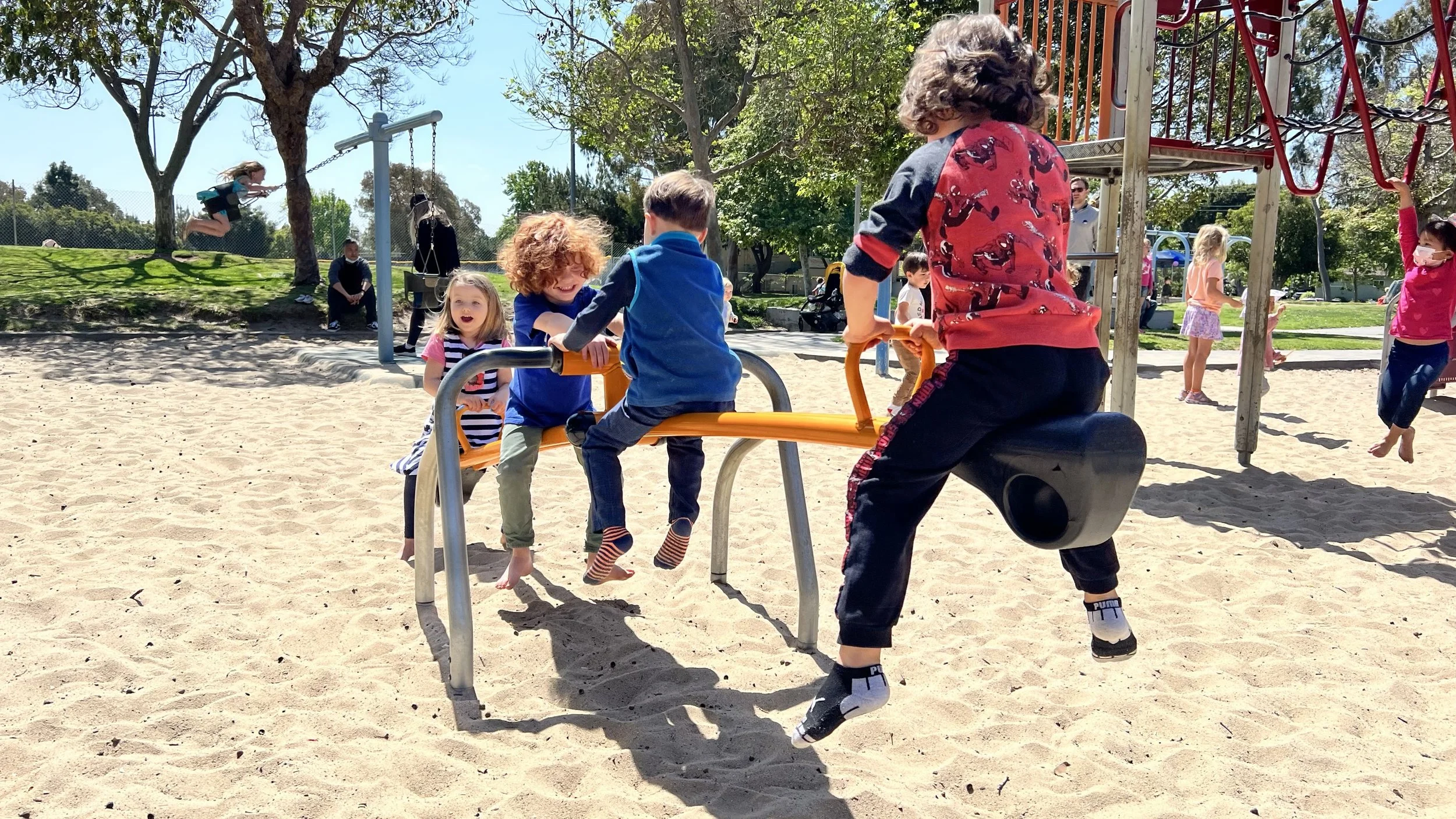 Children playing on a seesaw at a playground on a sunny day, with other kids on swings and climbing structures in the background.