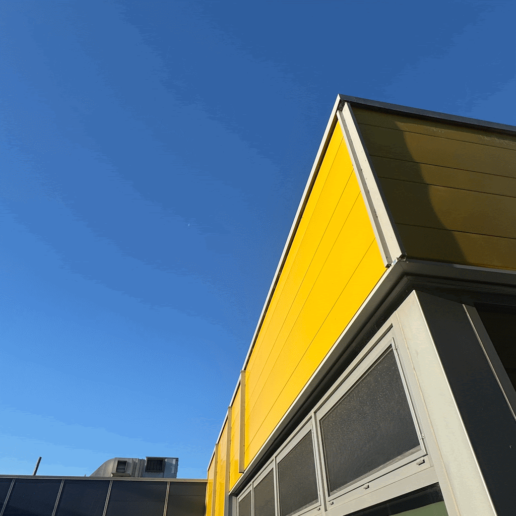 Close-up of the corner of a modern building with yellow siding and gray metal framing against a clear blue sky.