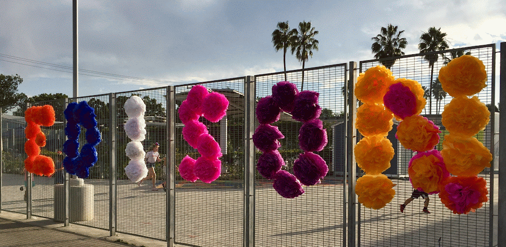 Colorful pom-pom decorations hanging on a metal fence at an outdoor sports court or playground, with palm trees and a cloudy sky in the background.
