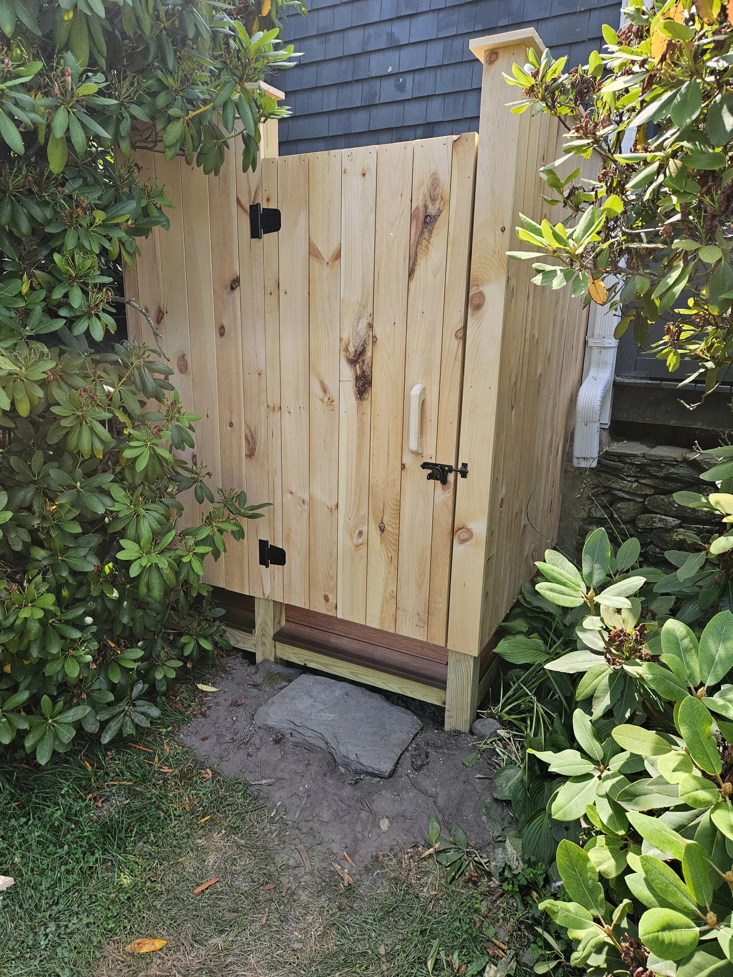 A full view of the outdoor shower shows its simple, clean-lined pine enclosure set against lush greenery near Ragged Island Brewery. The natural wood is sealed to enhance durability while preserving its warm, rustic tone. Trex decking forms the base,