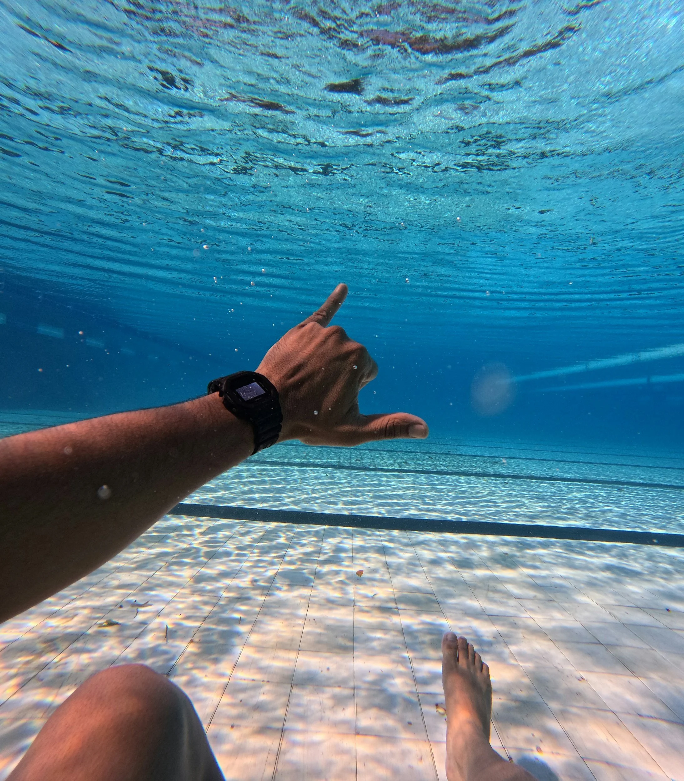 Underwater view of a person sitting at the edge of a swimming pool, with one hand extended and making a shaka sign.