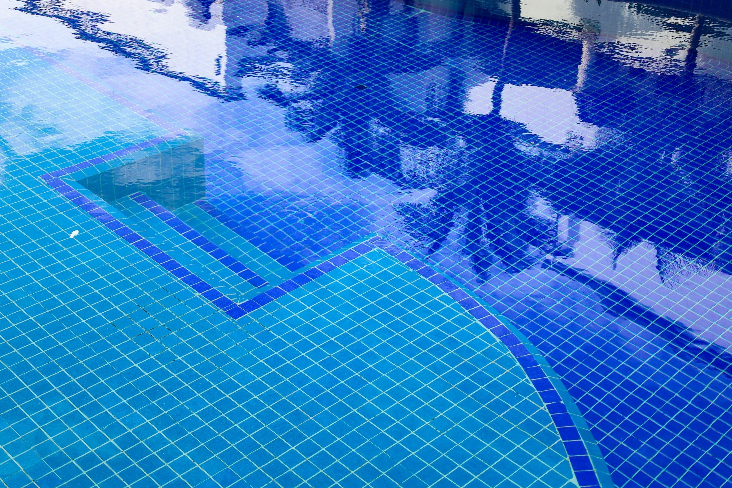 Reflection of palm trees and sky on the surface of a swimming pool with blue tiles.