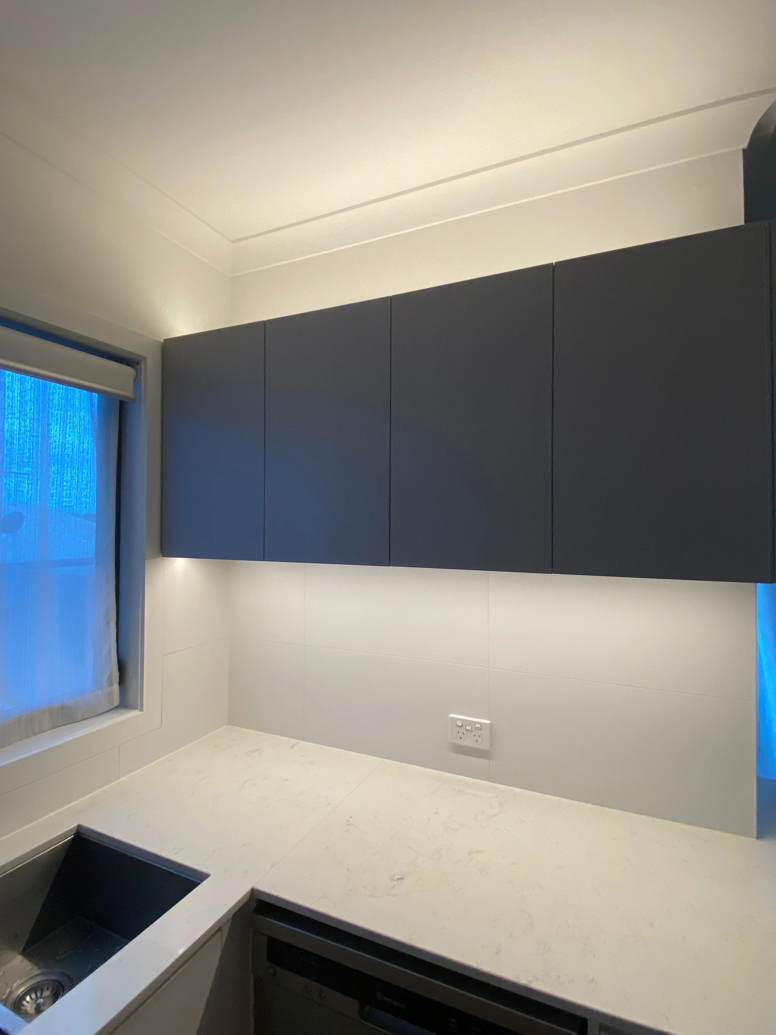 Kitchen corner with white marble countertop, dark gray upper cabinets, white wall tiles, a small window with blue light outside, and a double power outlet on the wall.