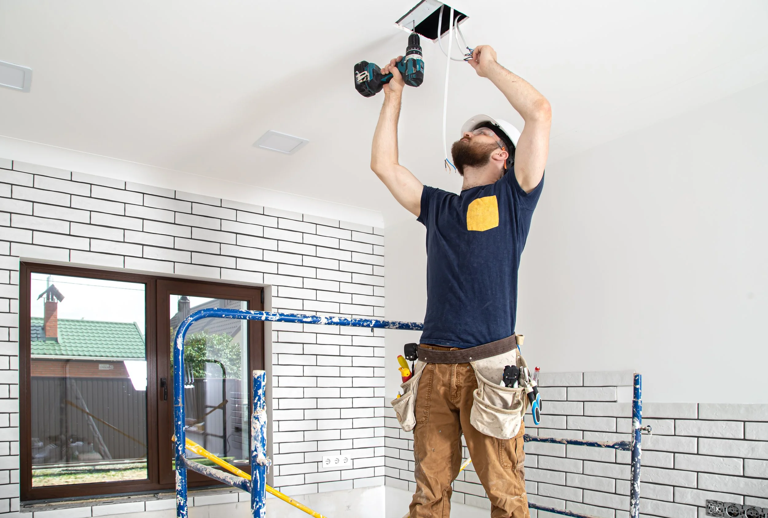 A man wearing a white hard hat and safety glasses installing a ceiling fixture with a power drill in a room with white brick walls and a window.