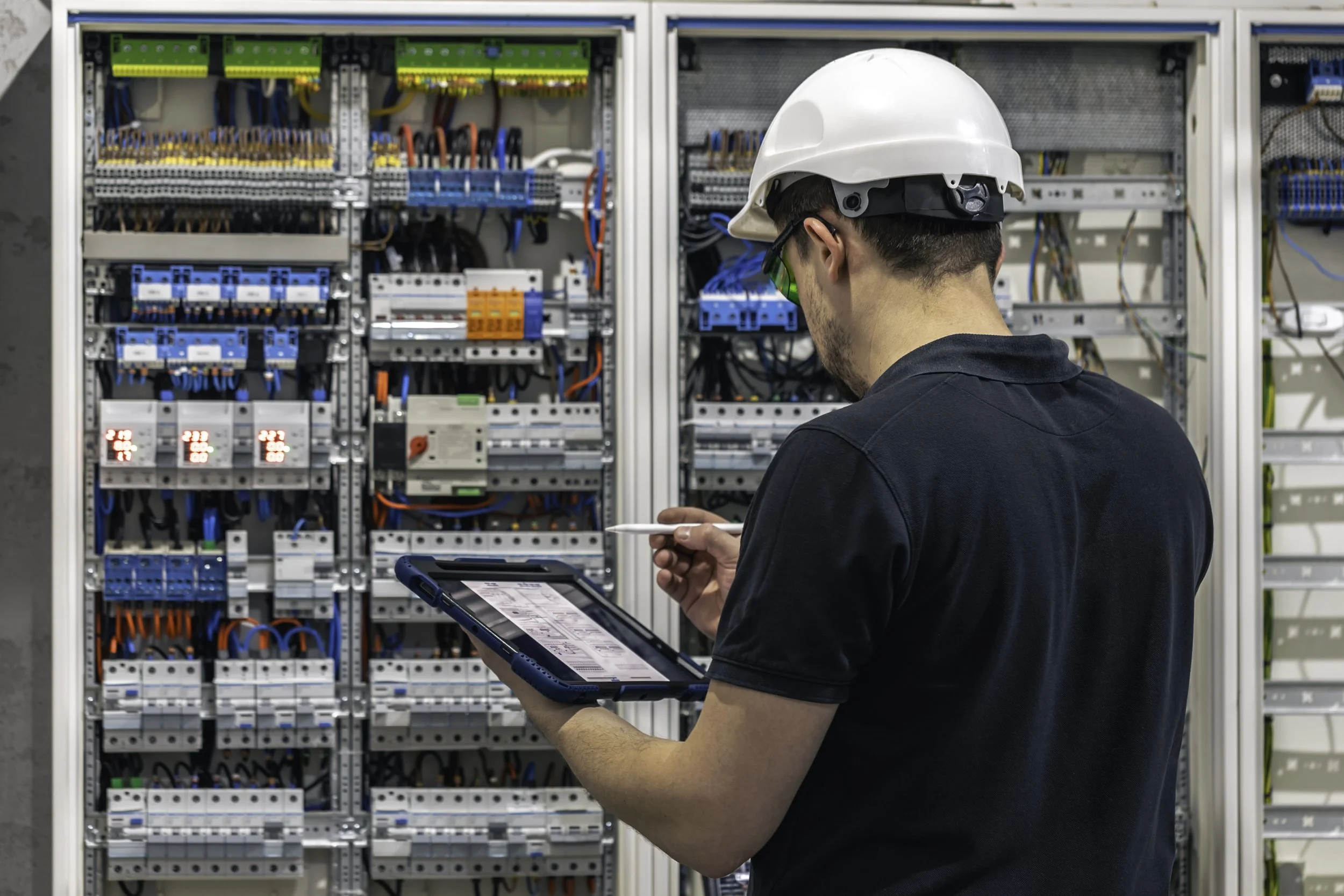 Technician in safety gear inspecting electrical control panel with tablet in hand.