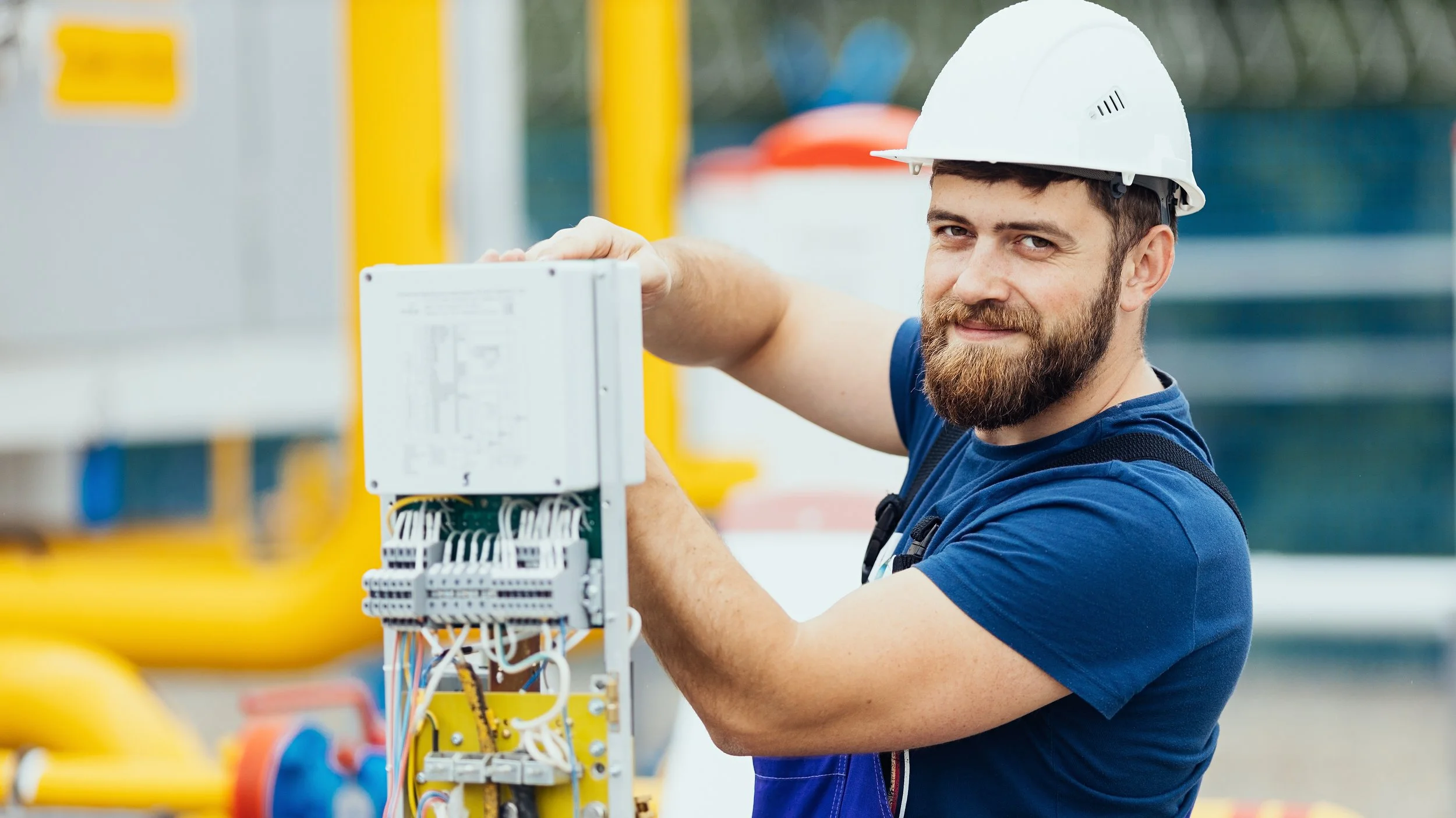 A male worker wearing a white safety helmet and blue shirt working on electrical equipment outdoors.