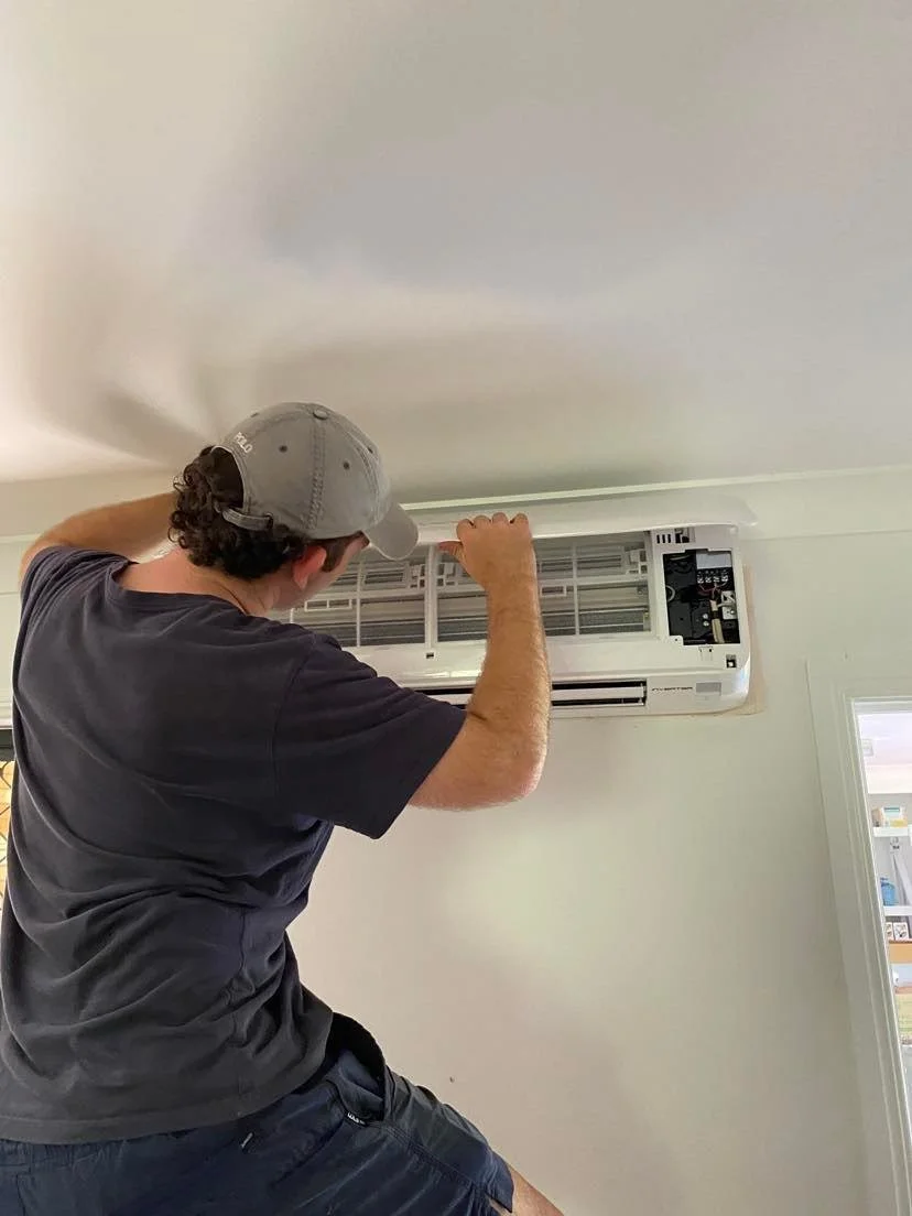 A man wearing a gray cap and black t-shirt is repairing or servicing a wall-mounted split air conditioning unit in a room.