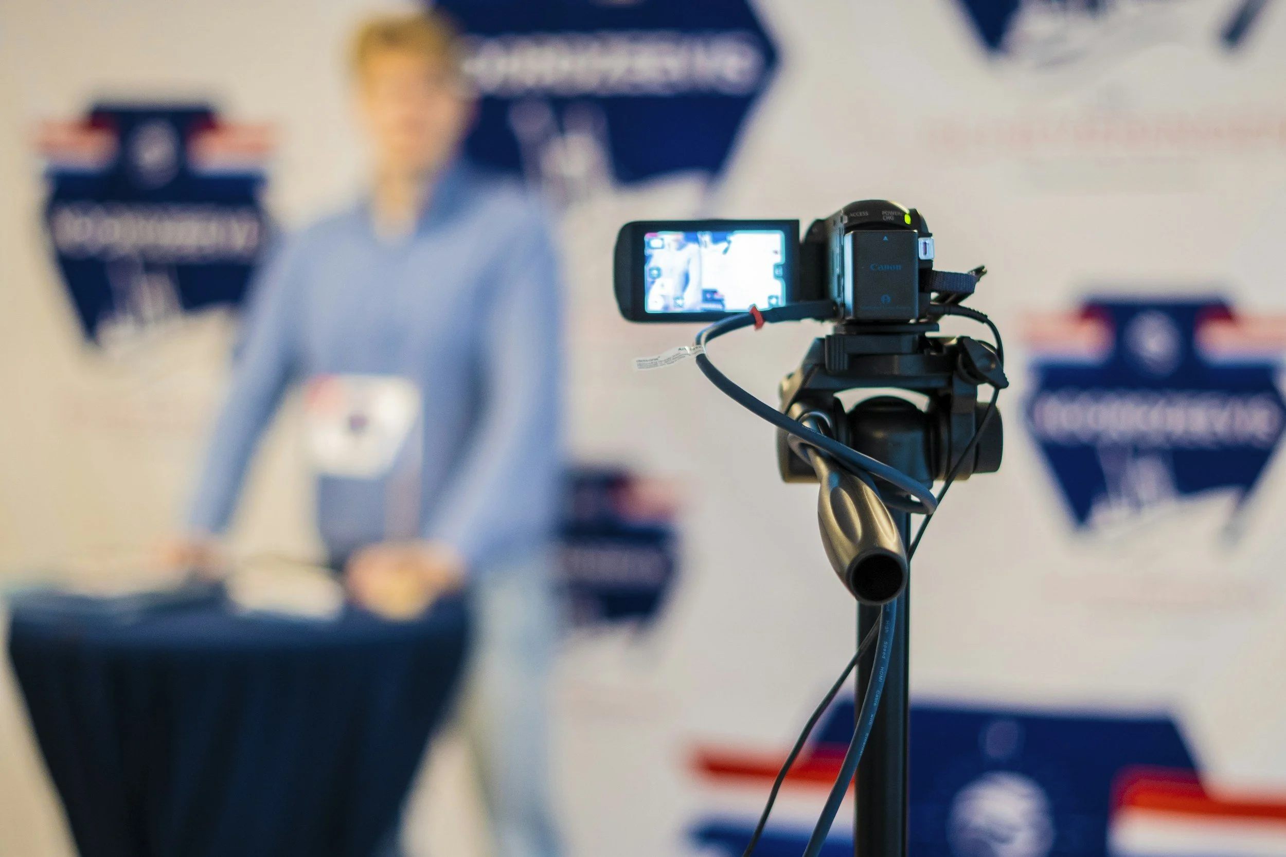 A professional video camera on a tripod pointing at a blurred woman speaking at a podium in front of a backdrop with logos.