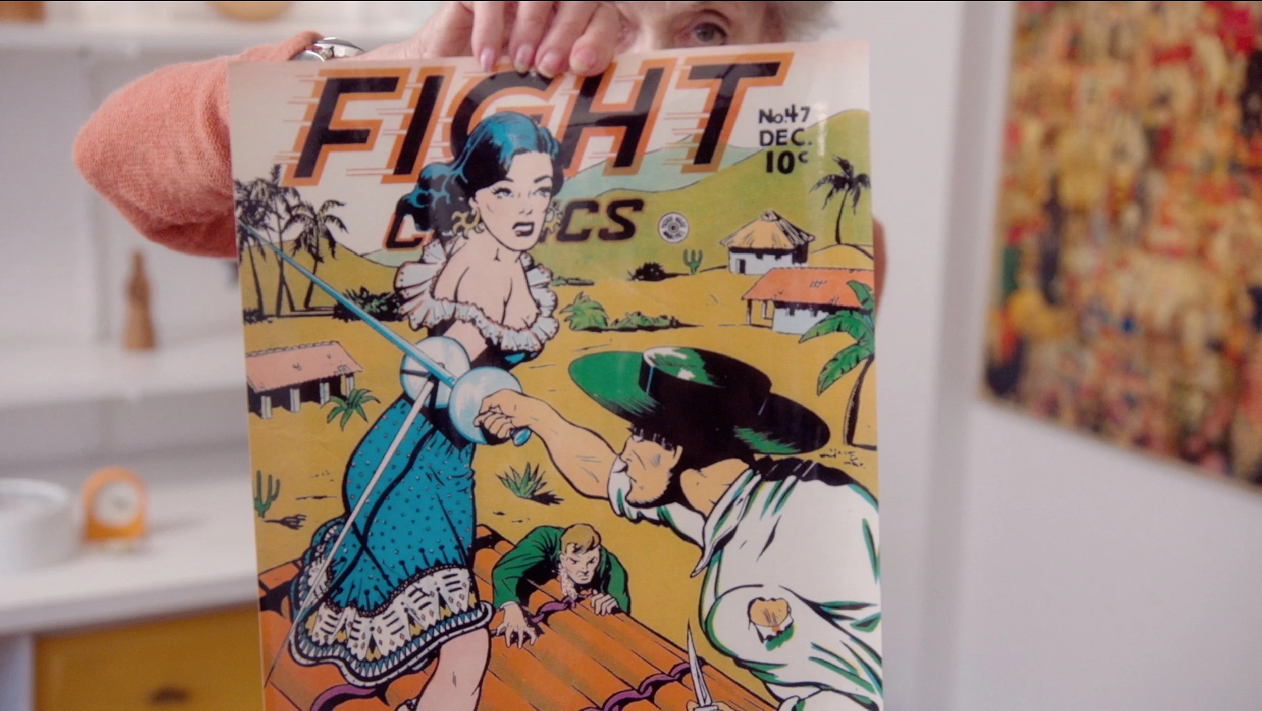 A person holds a comic book titled "FIGHT COMICS" with a colorful illustration of a woman with blue hair, a man in a sombrero, and a young boy on a tropical island.