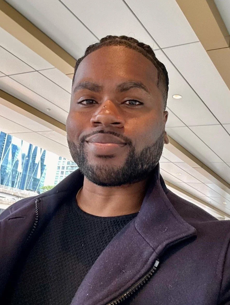 A man with a beard and braided hair smiling in an indoor setting with large windows showing tall buildings outside.