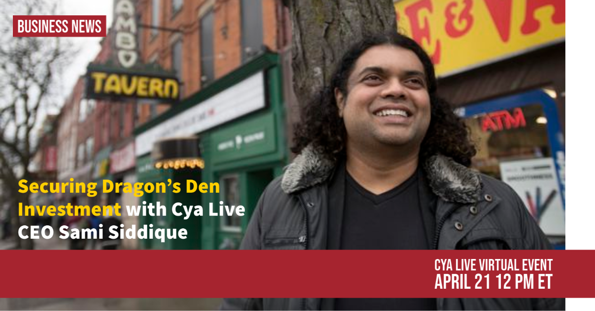 A man with long curly hair standing outside next to a tree, with storefronts and colorful signs in the background.