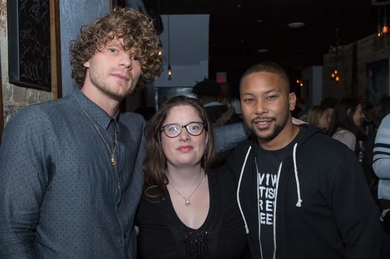 Three people standing together in a dimly lit bar or restaurant, smiling at the camera. The person on the left has curly hair, blue shirt, and light skin. The person in the middle has glasses, straight hair, and light skin. The person on the right ha