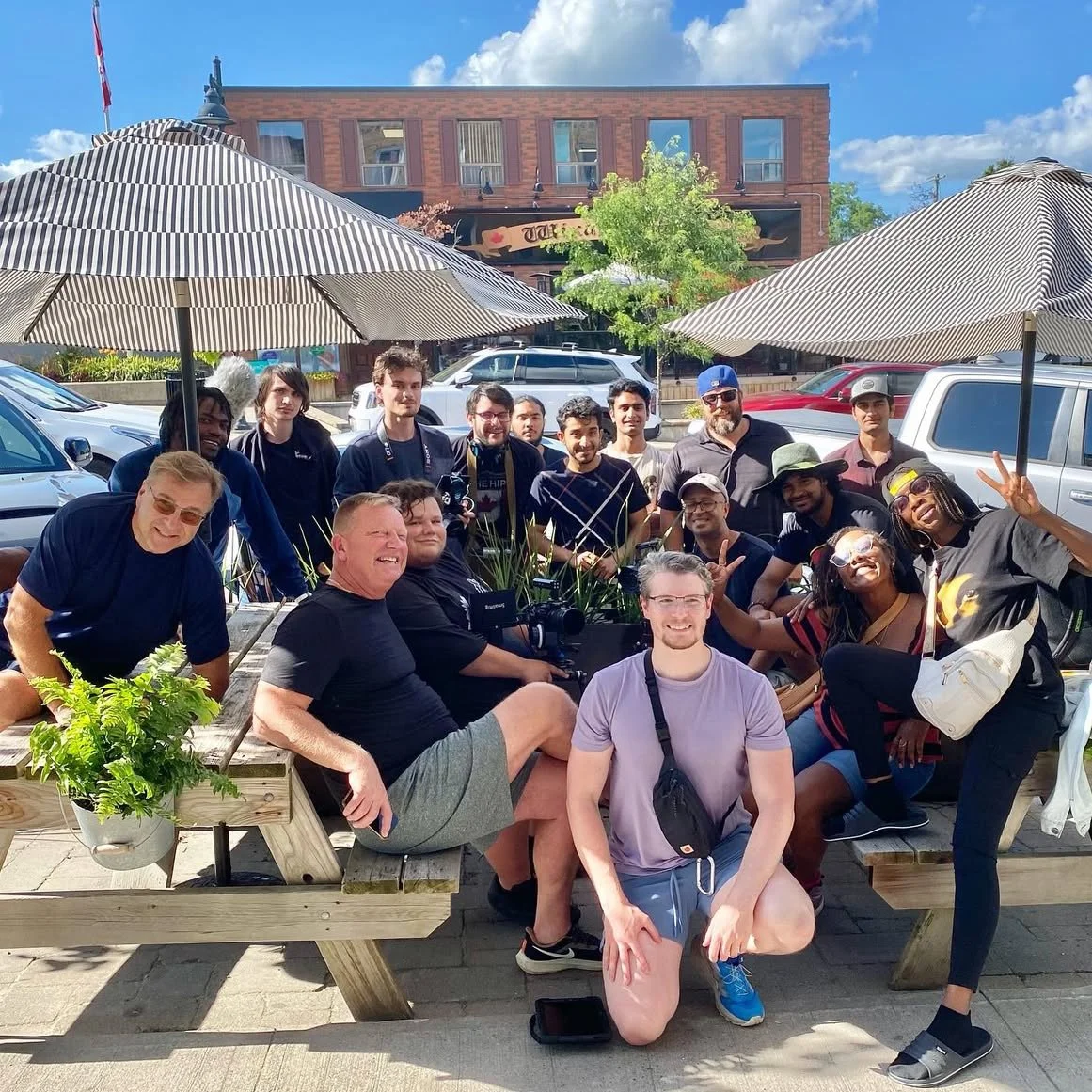 Group of people posing outdoors at a restaurant patio on a sunny day, with umbrellas overhead and a brick building in the background.