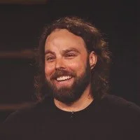 A man with long curly hair and a beard, smiling.