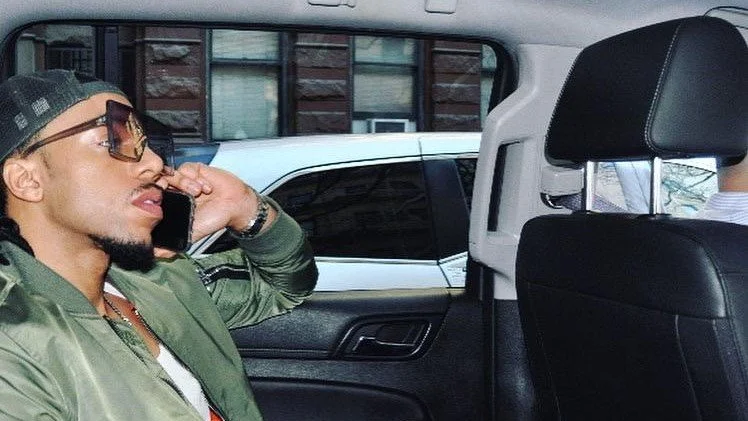 Man with sunglasses talking on a cellphone while sitting in the back seat of a car.