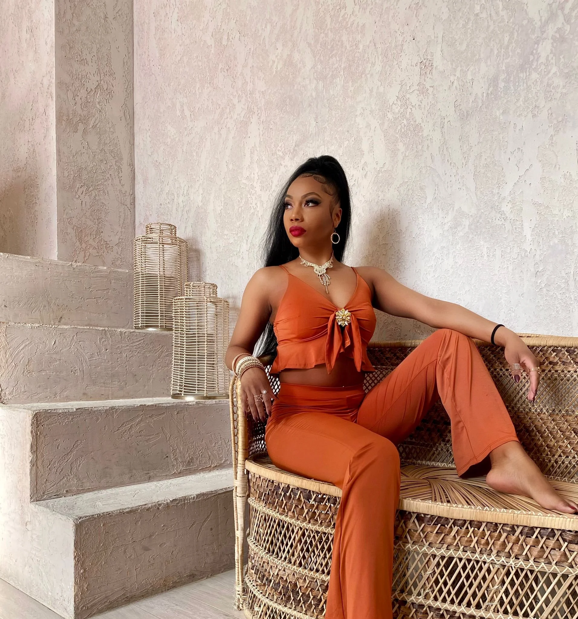 A woman in an orange two-piece outfit with jewelry, sitting on a wicker bench in a room with cream textured walls and decorative vases.