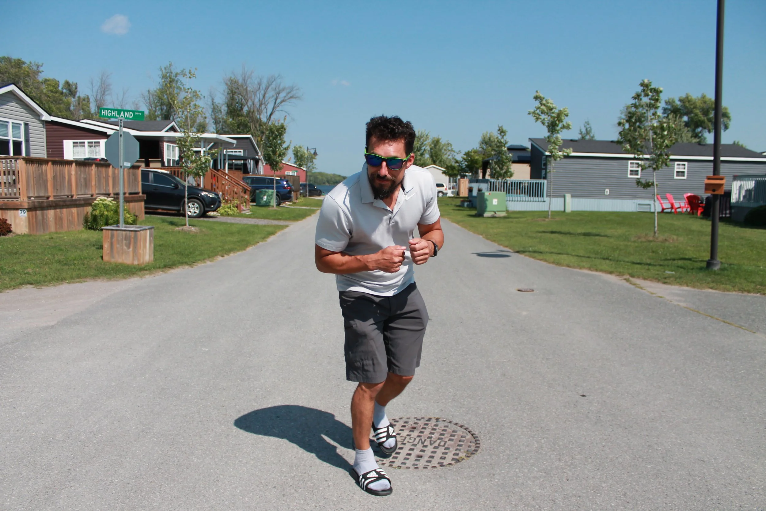 A man running on a suburban street wearing sunglasses, a white polo shirt, gray shorts, and sandals with socks.