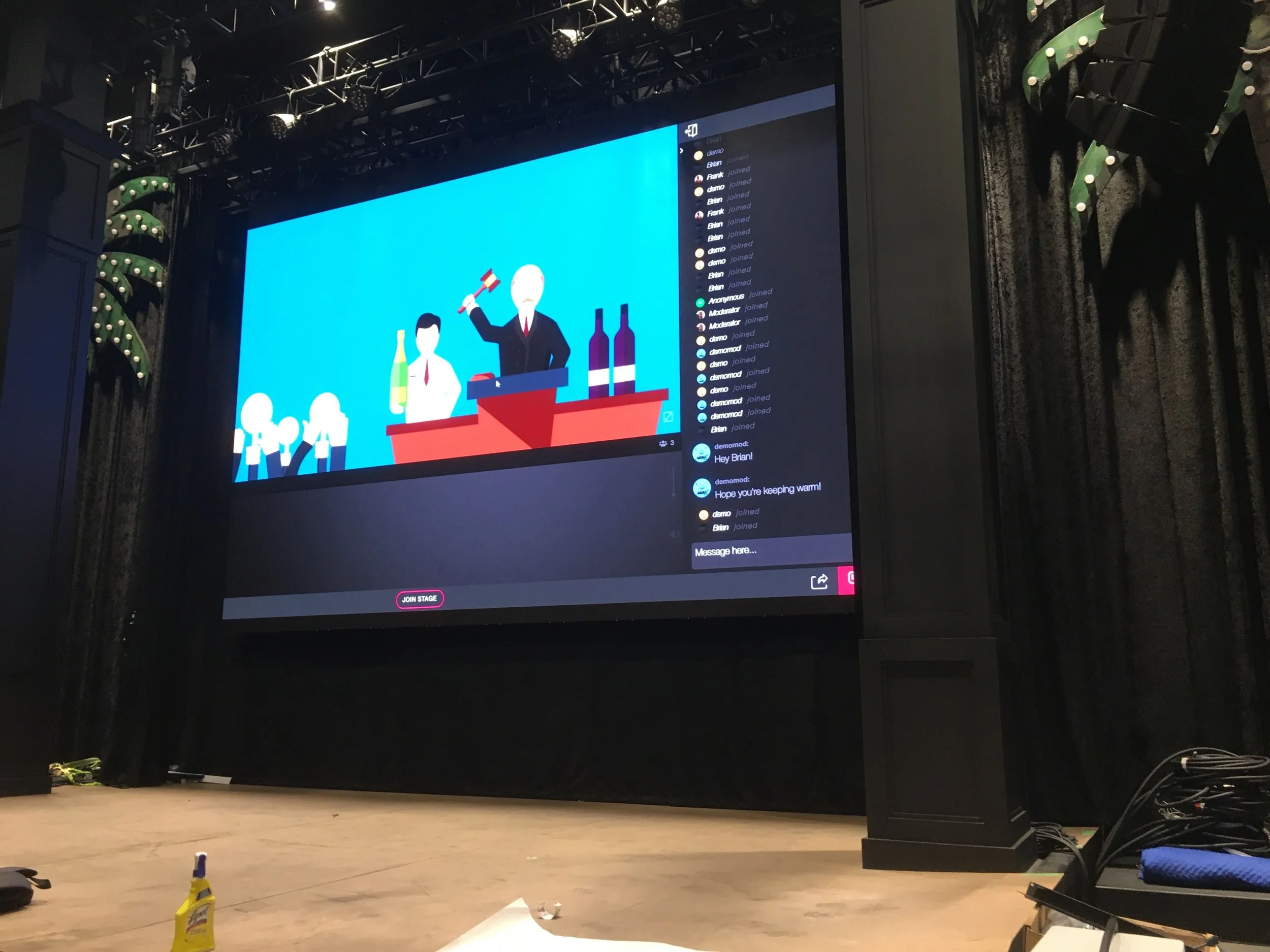 Large digital screen on stage showing animated illustration of people at a bar with bottles, and a man in a suit holding a hammer. The right side of the screen displays a chat window with user comments and joined notifications. The stage has black cu