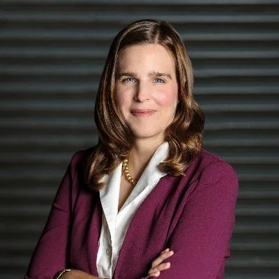 A woman with shoulder-length brown hair wearing a white blouse and a maroon blazer posing with arms crossed in front of a dark, striped background.