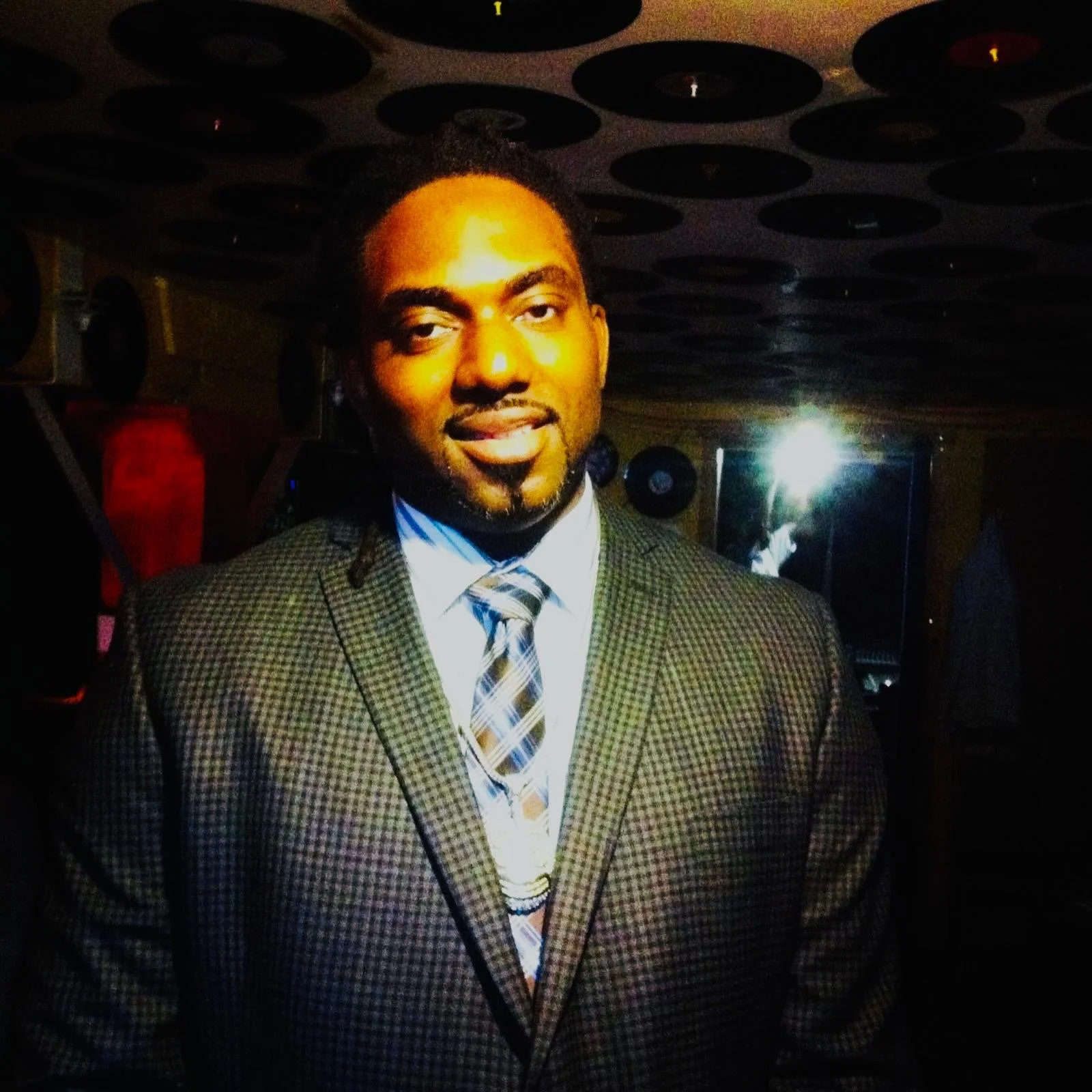 A man wearing a checkered suit, white shirt, and a plaid tie, smiling in a dimly lit room with vinyl records on the ceiling and a mirror reflecting a person playing guitar in the background.