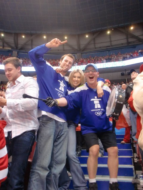 Three people celebrating at a sports event, with two wearing Vancouver 2010 Olympic jerseys and one holding a drum.
