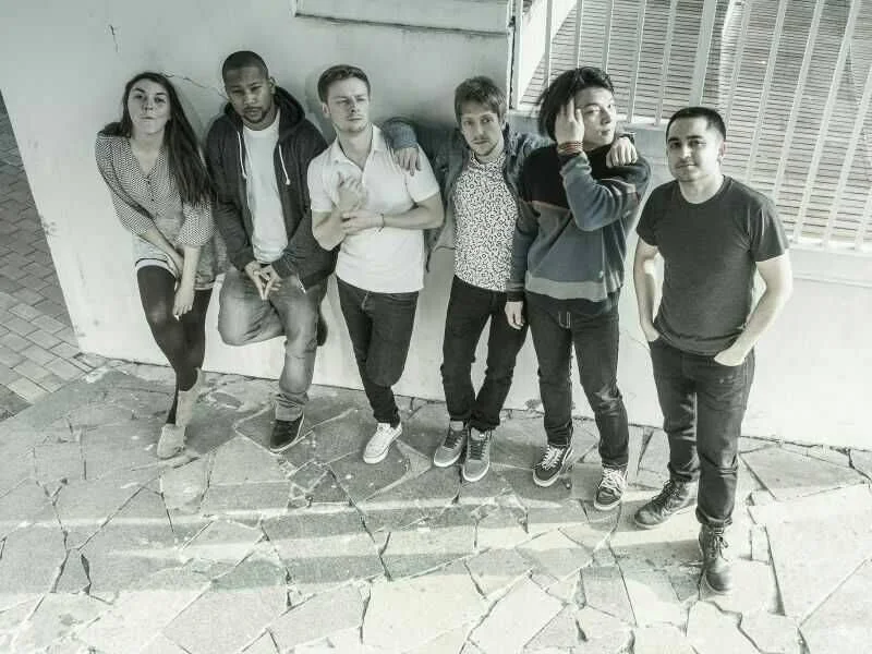 Group of six young people standing together outdoors near a wall with metal bars, with cracked pavement in the foreground.