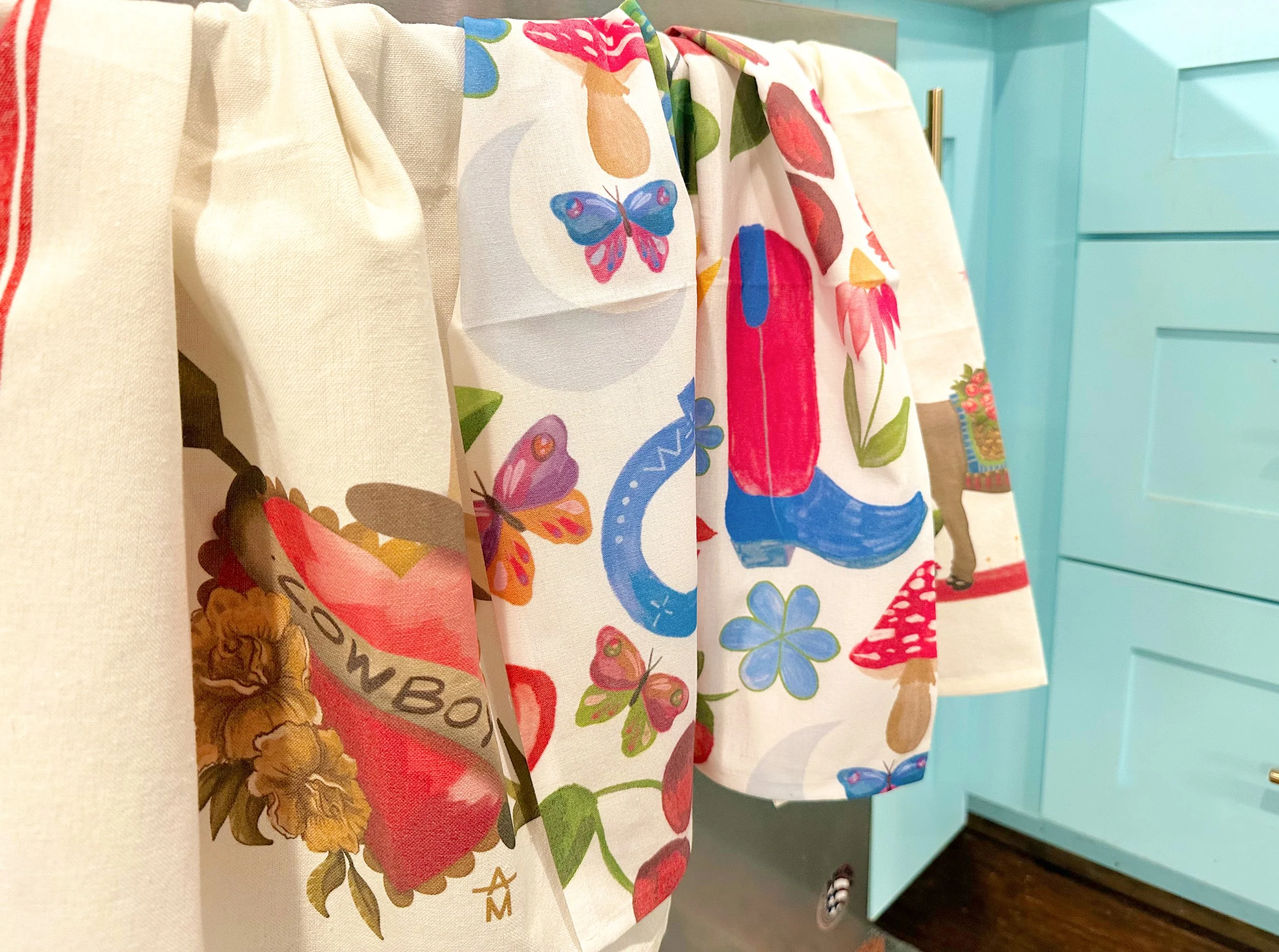 Colorful kitchen towels featuring various designs, hanging in front of light blue cabinets.