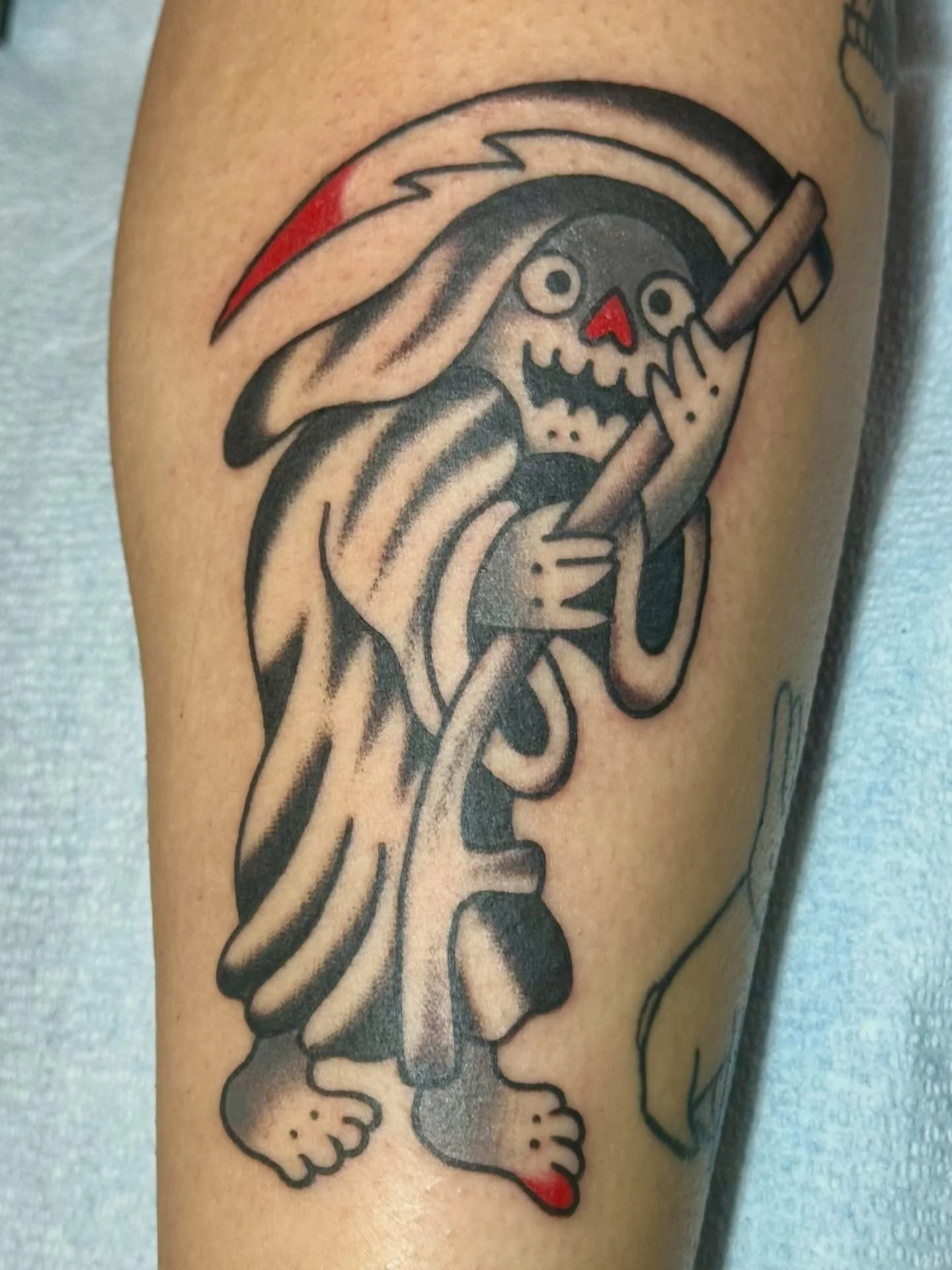 Tattoo of the Grim Reaper with a red nose and a skull face, holding a stick, with a red-tipped scythe on its head.