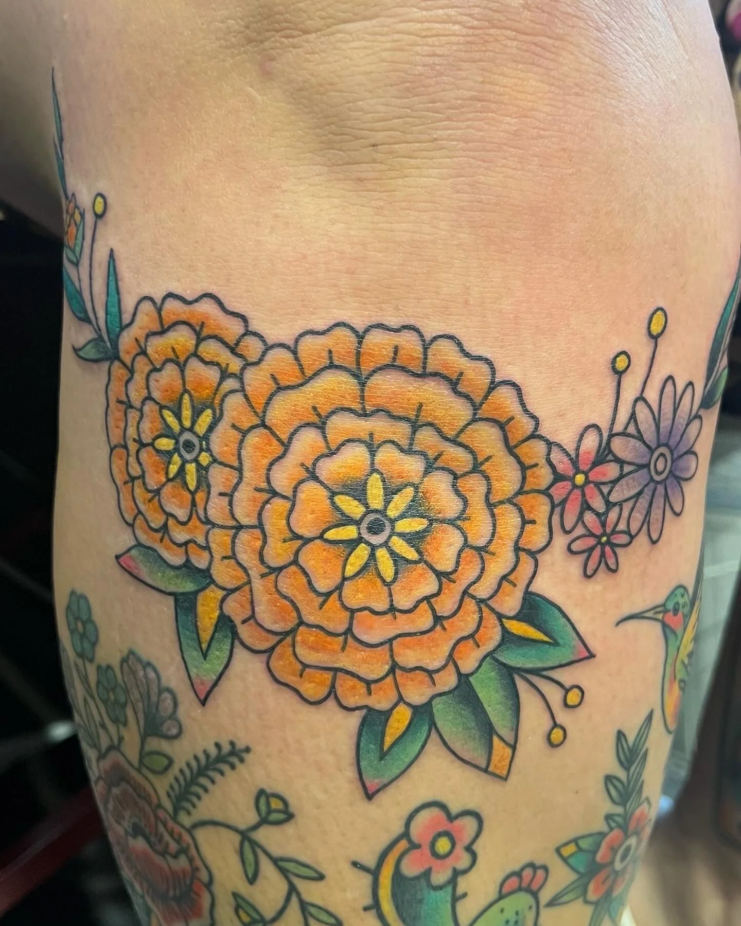 Colorful tattoo of orange and yellow marigold with green leaves and a hummingbird on a person's skin.