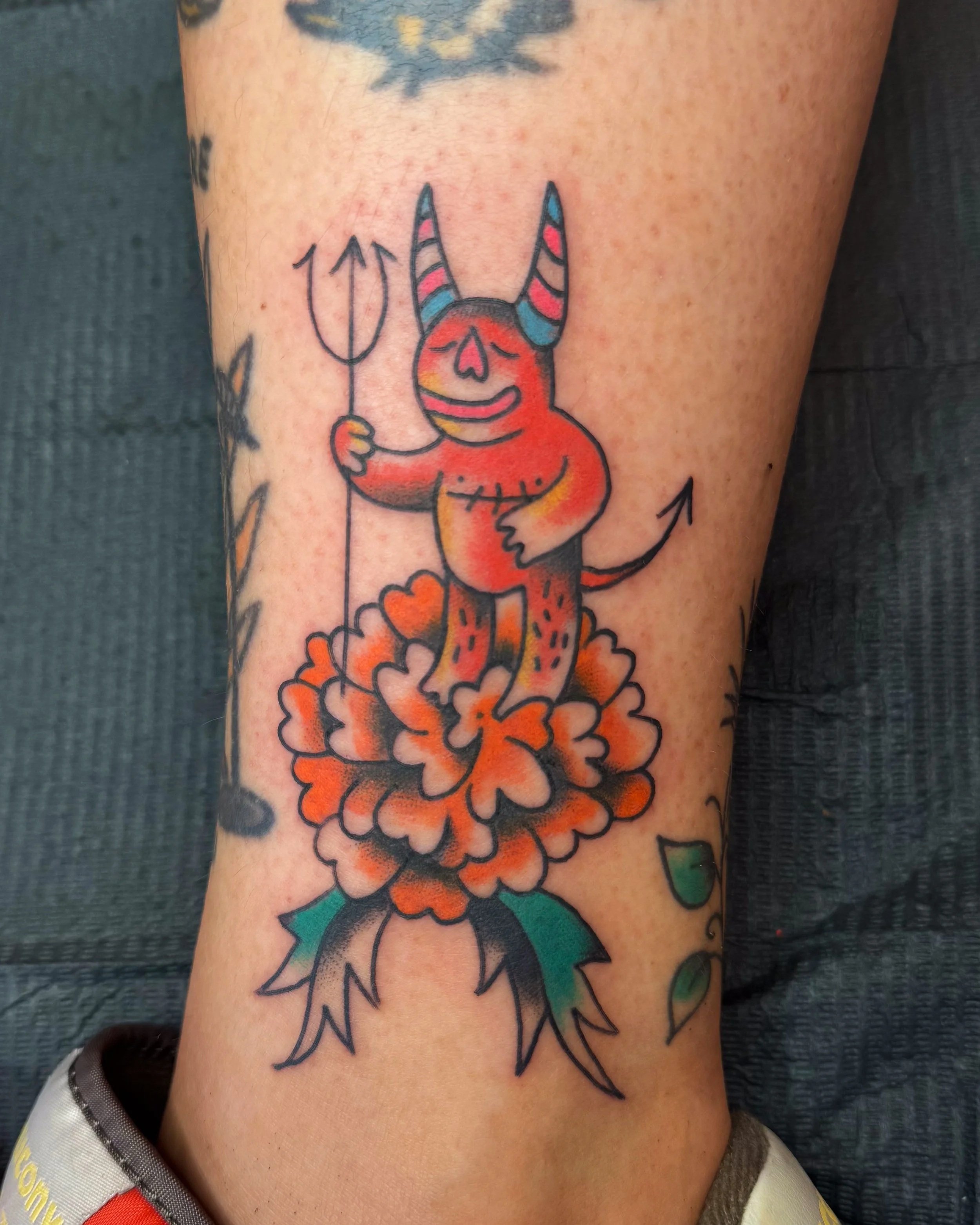 Tattoo of a cartoon devil standing on a flower, holding a trident, with a smiling expression.