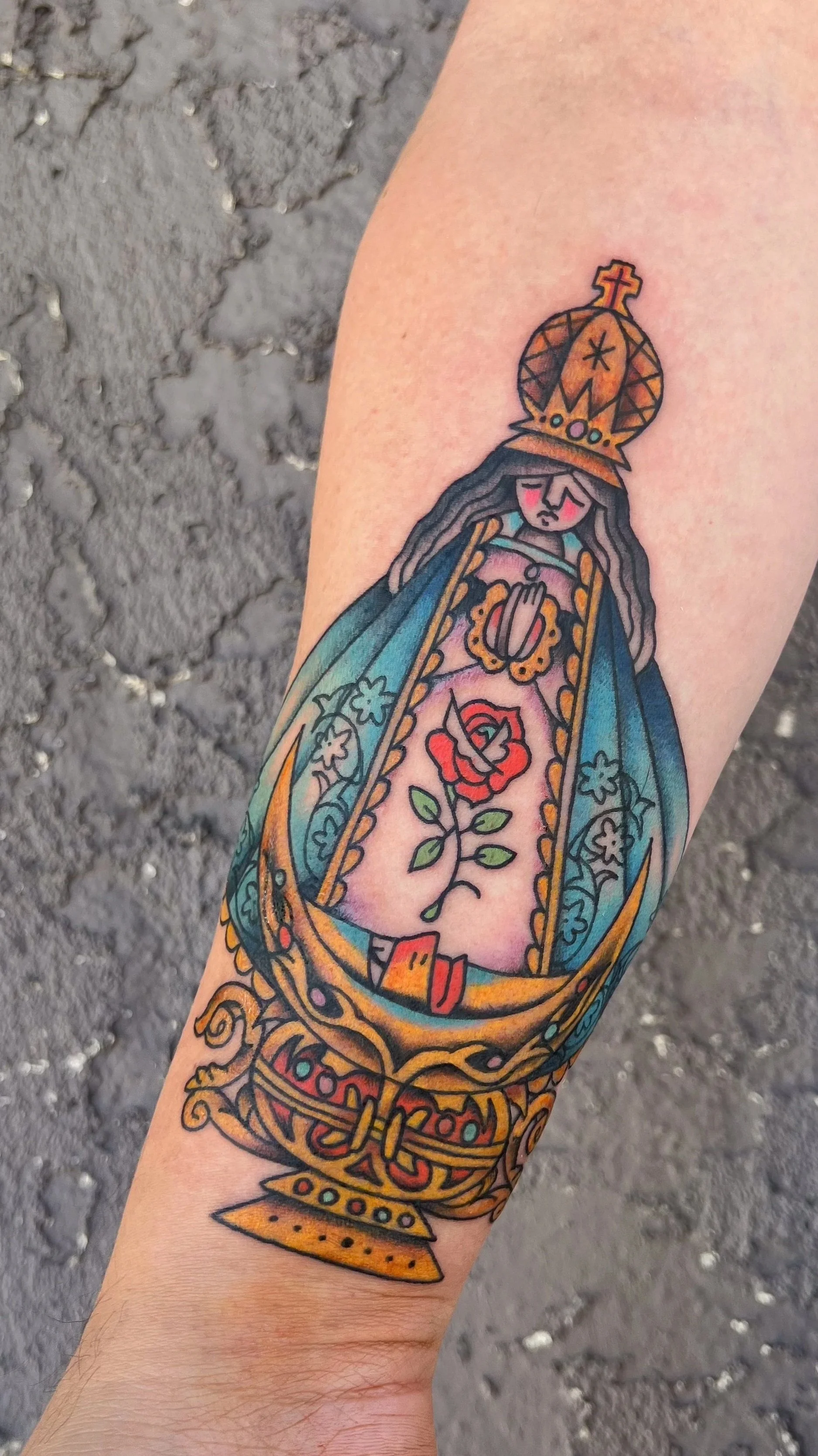 Colorful tattoo of a depiction of Our Lady of Guadalupe on a person's arm, with traditional Mexican religious iconography and floral details.